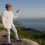 Kabir is wearing a white shirt, pale blue jeans and a light brown fedora hat. He is standing high up on a hillside, sideways to the camera, looking out to and gesturing to sea.